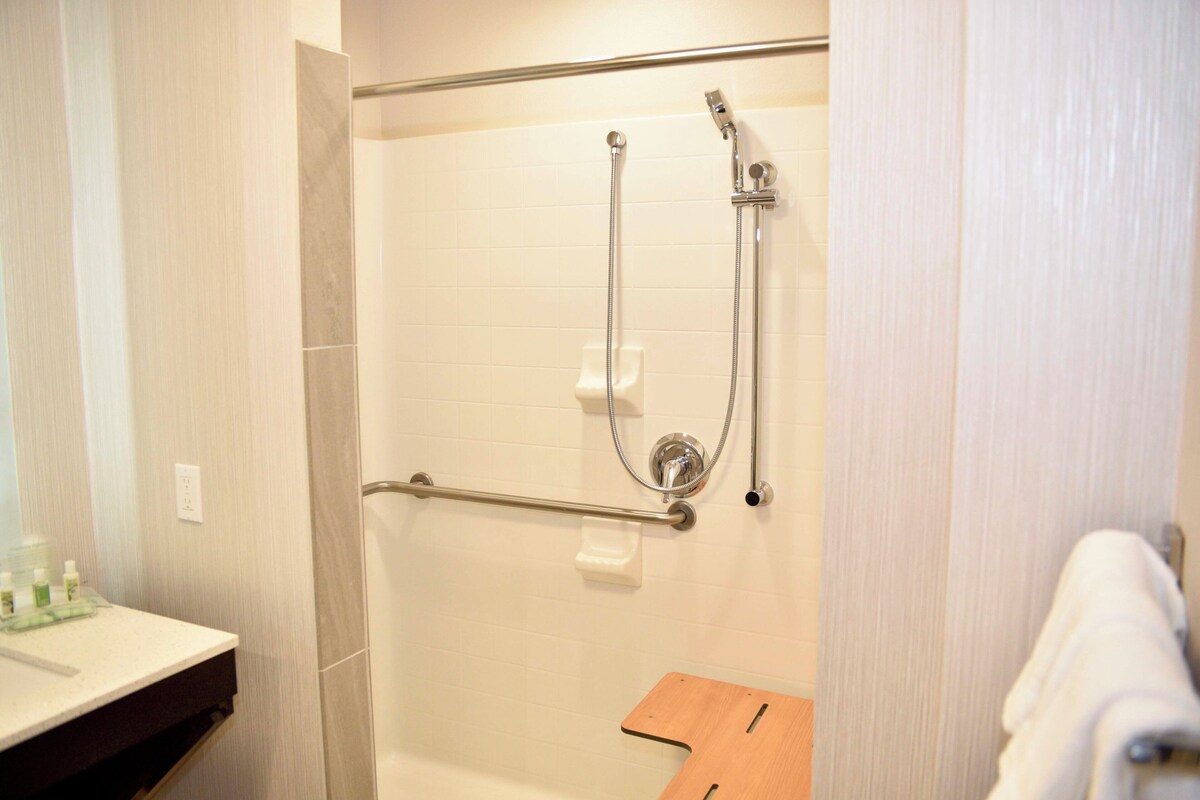 Een douche met een handgreep, een witte betegelde muur en een houten bankje.