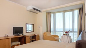Una habitación de hotel con cama, escritorio con televisor, silla, mesita con un florero y ventana con cortinas.