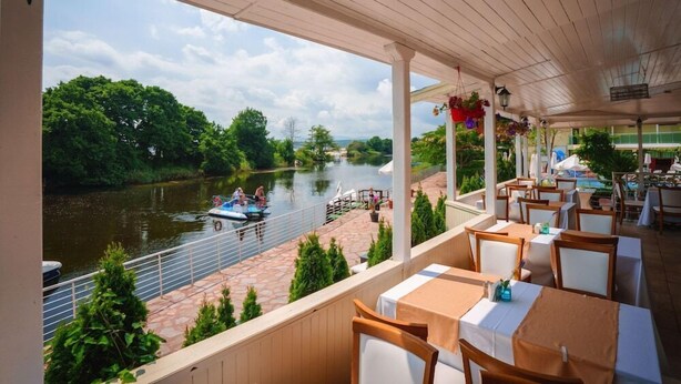 Un área de comedor al aire libre techada, con mesas y sillas, con vista a un río con barcos y vegetación.