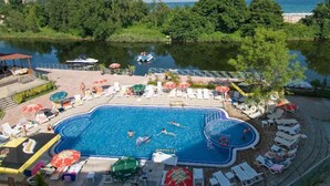 Una piscina con sillas de descanso y sombrillas, rodeada por un lago y árboles.