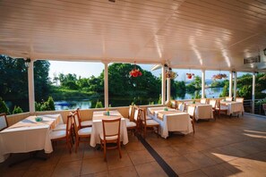 A covered outdoor dining area with tables and chairs, large windows overlooking a lake, and hanging flower baskets. - Perla Sun Park Hotel (Primorsko)