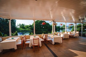 Un área de comedor al aire libre con techado, mesas y sillas, amplias ventanas con vista a un lago, y cestas de flores colgantes.
