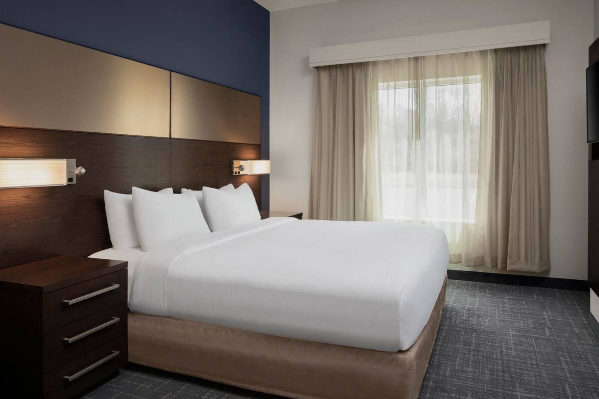 Een hotelkamer met een groot bed, een houten hoofdbord, een nachtkastje met lades, en een raam met dunne gordijnen.