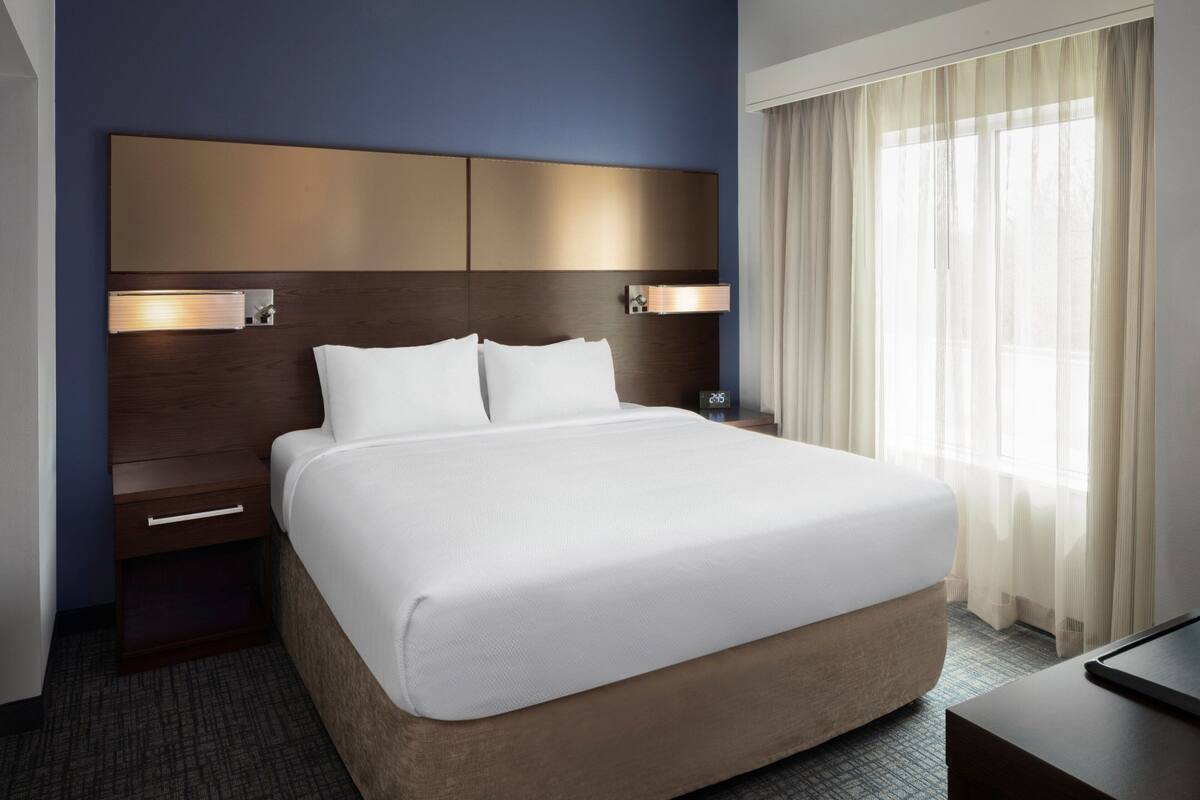 Een hotelkamer met een groot bed, een houten hoofdbord, nachtlampjes, een nachtkastje en een raam met dunne gordijnen.
