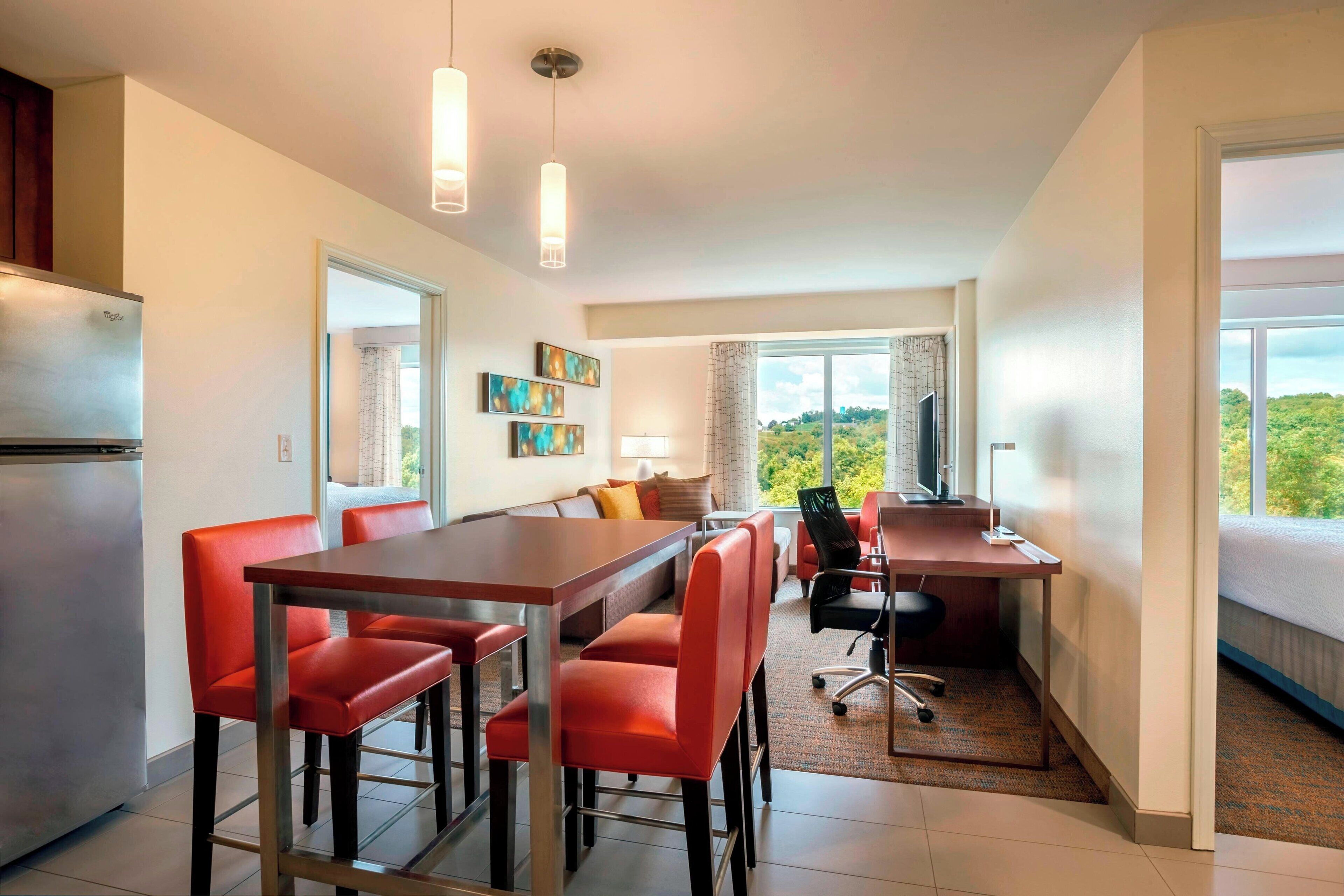 A modern kitchen with a dining area, a workspace, and a large window with a view of greenery.