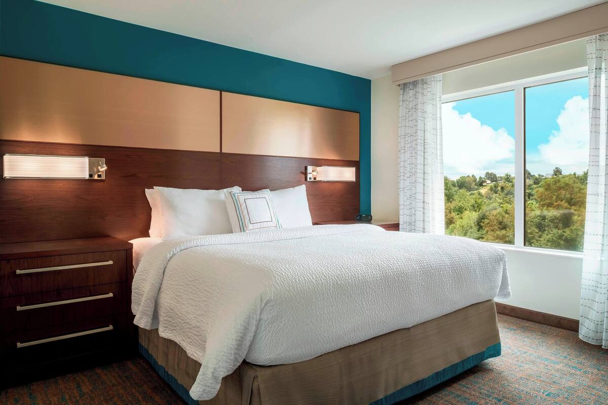 Een hotelkamer met een groot bed, een houten hoofdbord, een nachtkastje en een raam met uitzicht op groen.