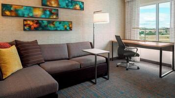 A modern hotel room with a sectional sofa, a desk with a chair, a standing lamp, and abstract wall art.