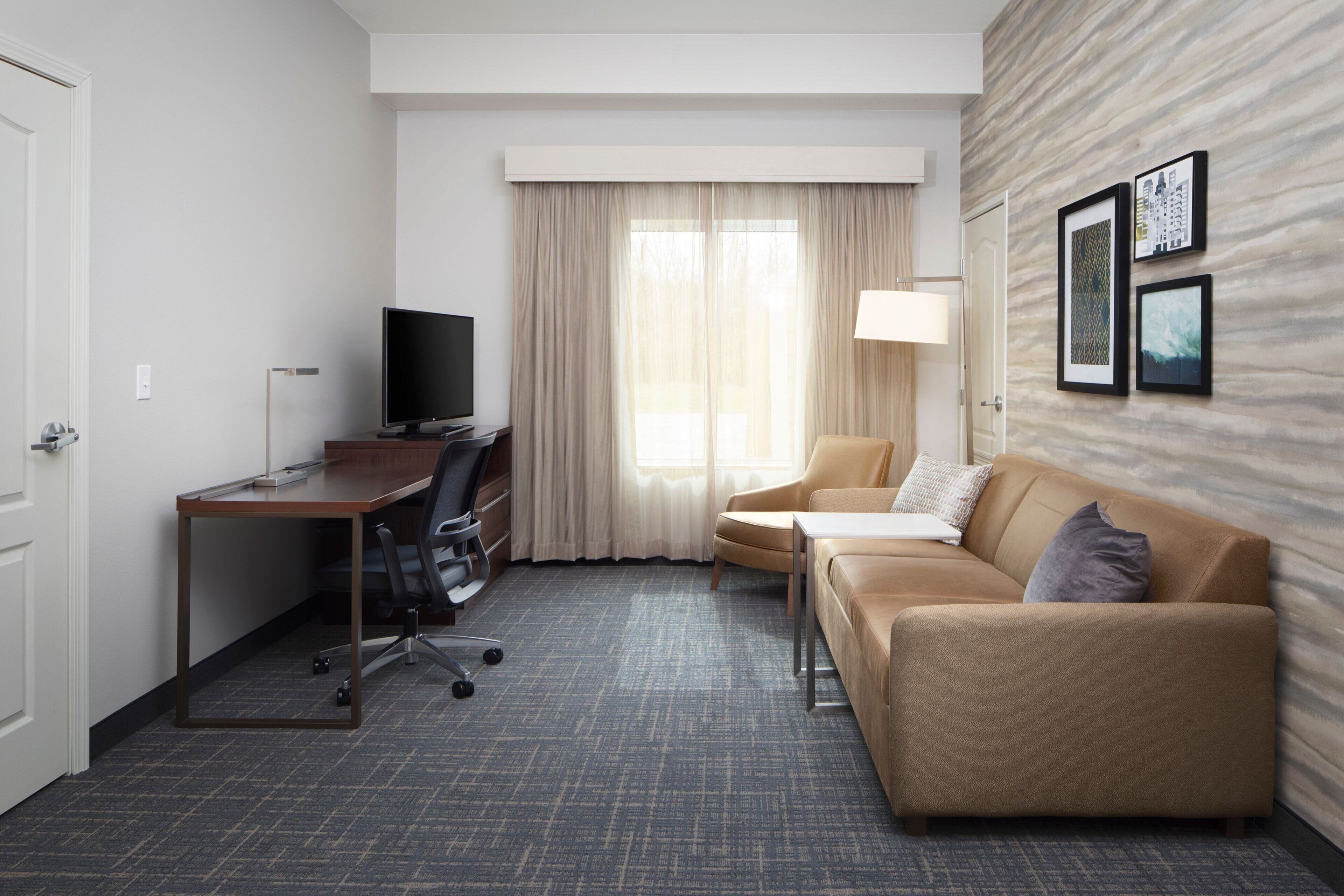 Quarto de hotel com escrivaninha, cadeira, sofá e televisão.