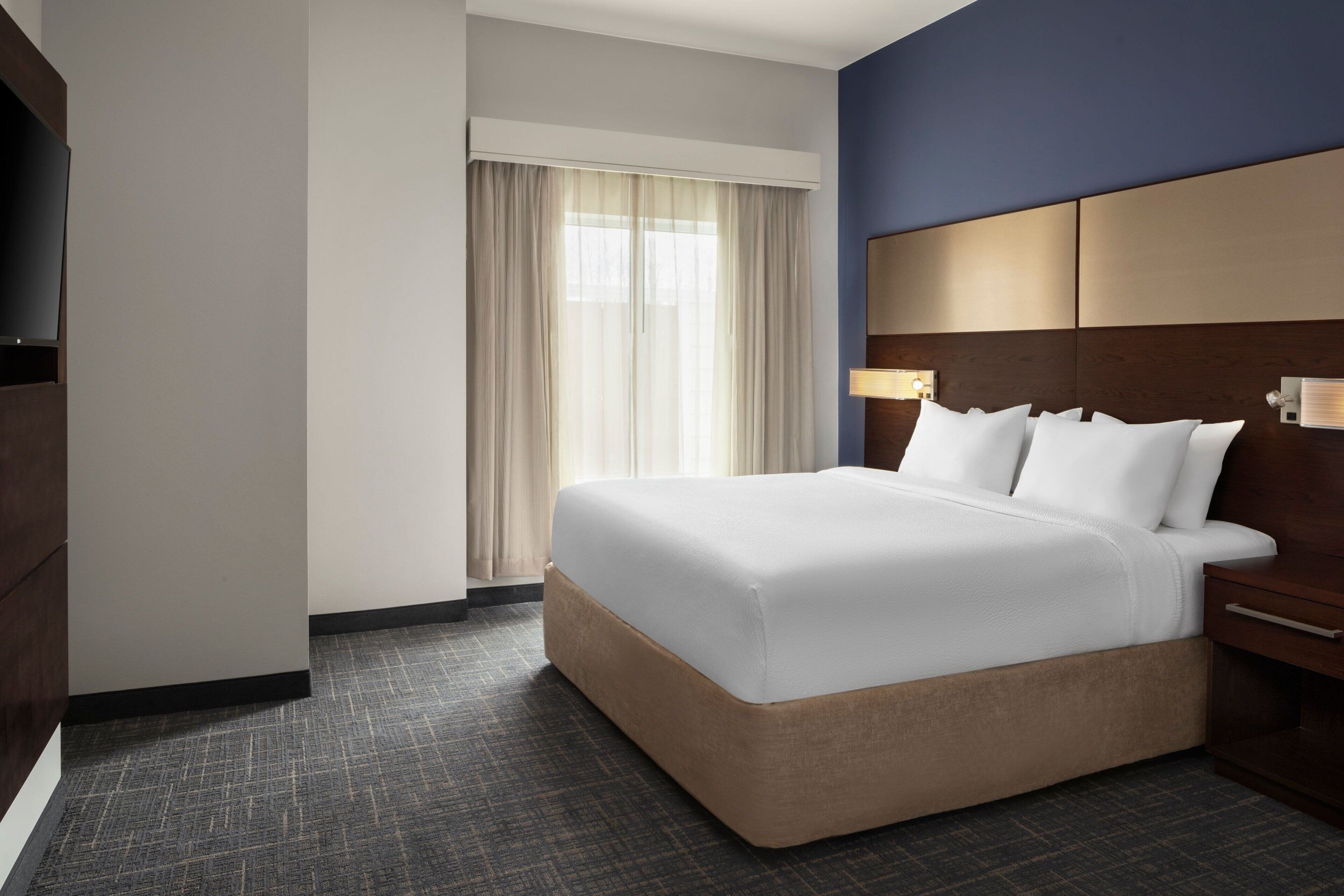 Habitación de hotel con una cama grande, cabecero de madera, televisor y ventana con cortinas.
