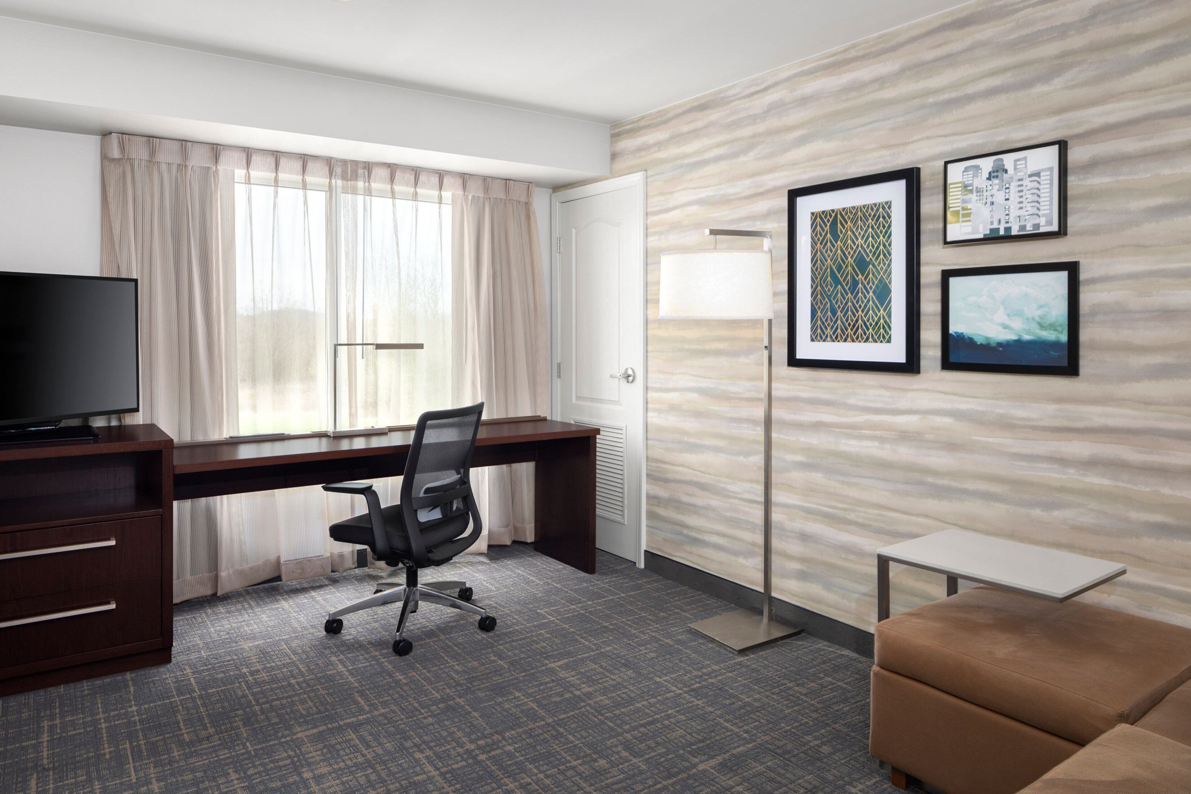 A hotel room with a desk, chair, television, sofa, and artwork on the wall.