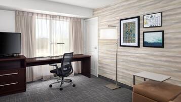 A hotel room with a desk, chair, television, sofa, and artwork on the wall.
