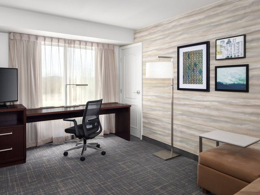 A hotel room with a desk, chair, television, sofa, and artwork on the wall.