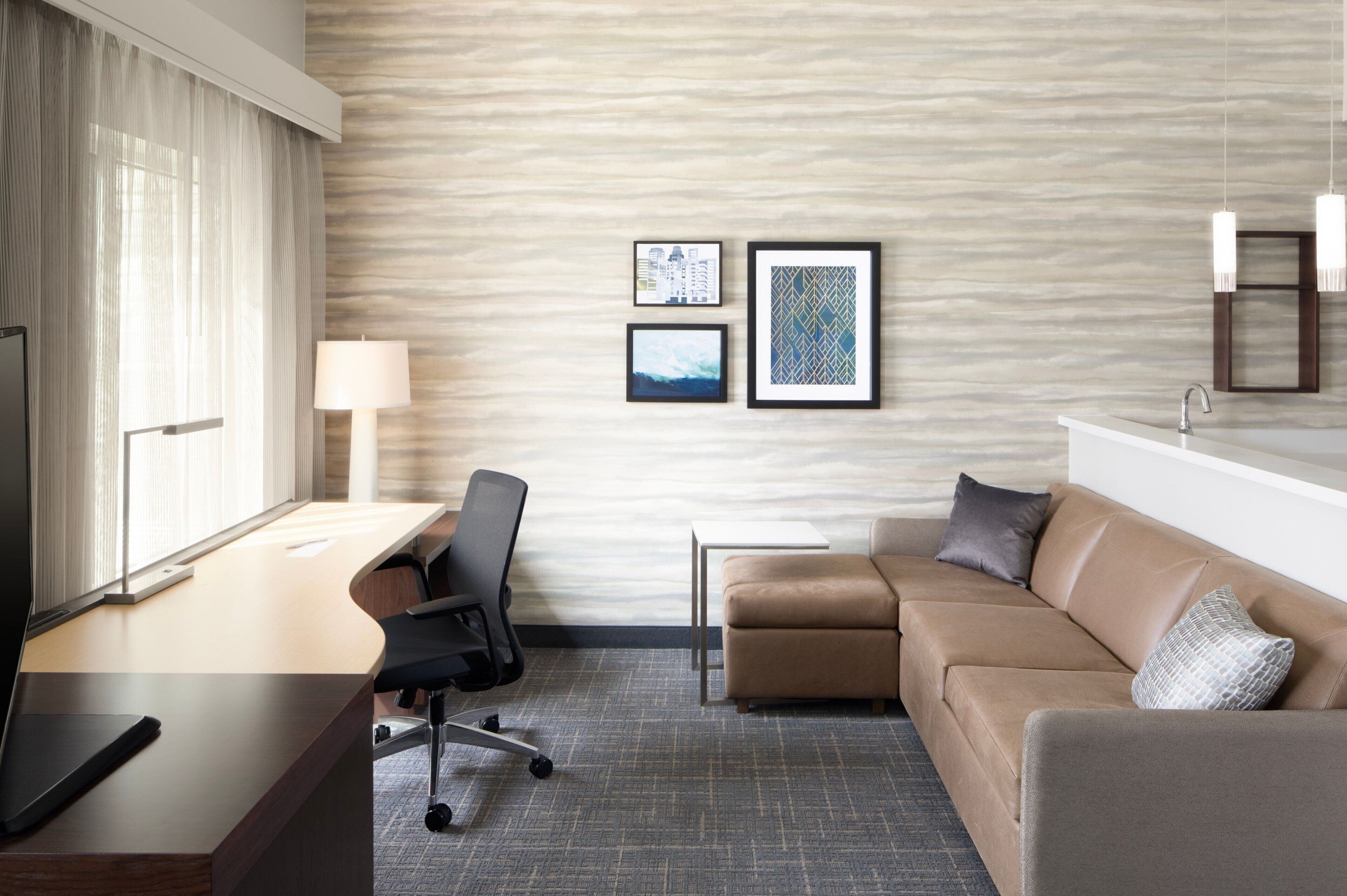 A hotel room with a desk, chair, sofa, and a bed. There are framed pictures on the wall, a lamp, and a TV.