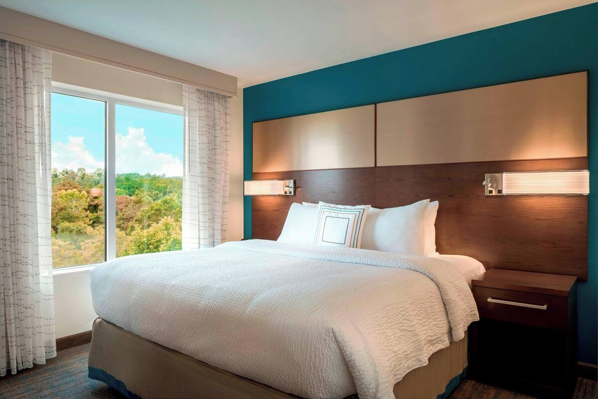 Een hotelkamer met een groot bed, een nachtkastje, een raam met gordijnen en uitzicht op bomen.