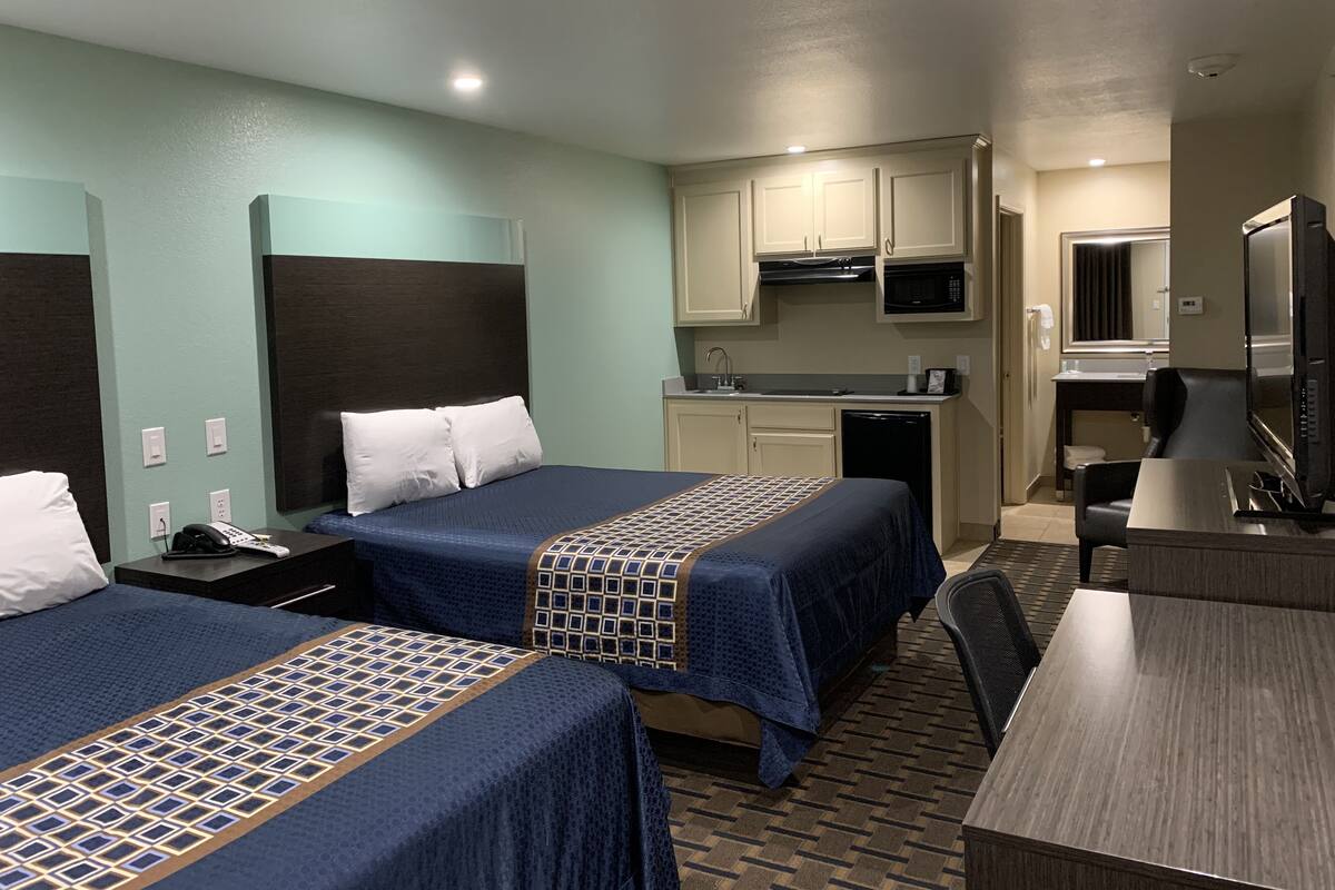 A hotel room with two beds, a kitchenette, a TV, and a desk.