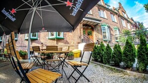 An outdoor seating area with wooden tables and chairs, a large black umbrella with a star logo, and a brick building in the background. - Hotel Logos (Gdansk)