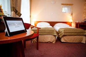 A hotel room with two beds, a bedside table with a tablet displaying "HOTEL LOGOS GDAŃSK", a red chair, and a lamp on the nightstand. - Hotel Logos (Gdansk)