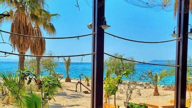 Uma área de estar ao ar livre com mesas e cadeiras, com vista para uma praia com palmeiras e o mar.