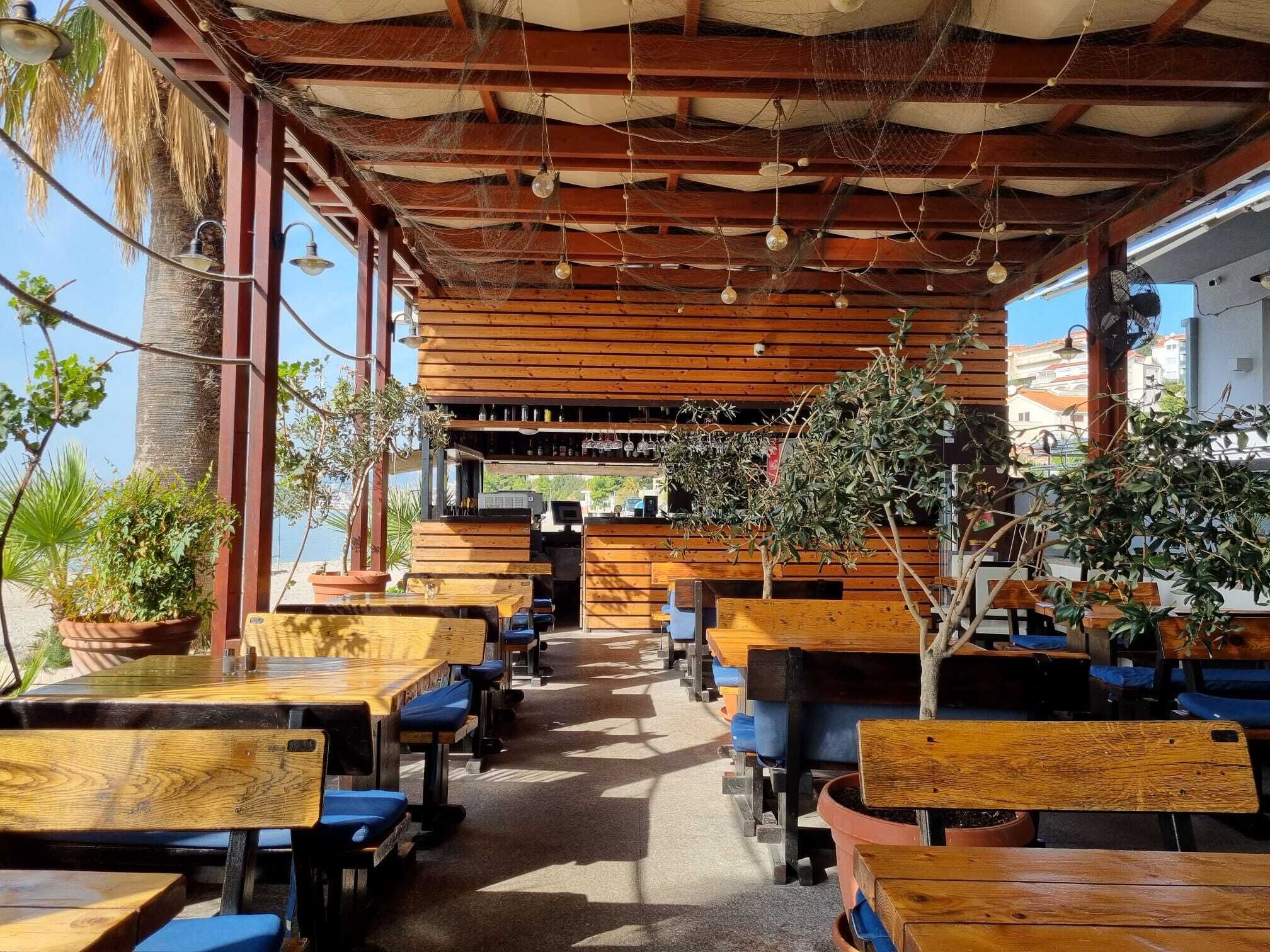 Un café al aire libre con mesas y sillas de madera, plantas en macetas y vista al mar.