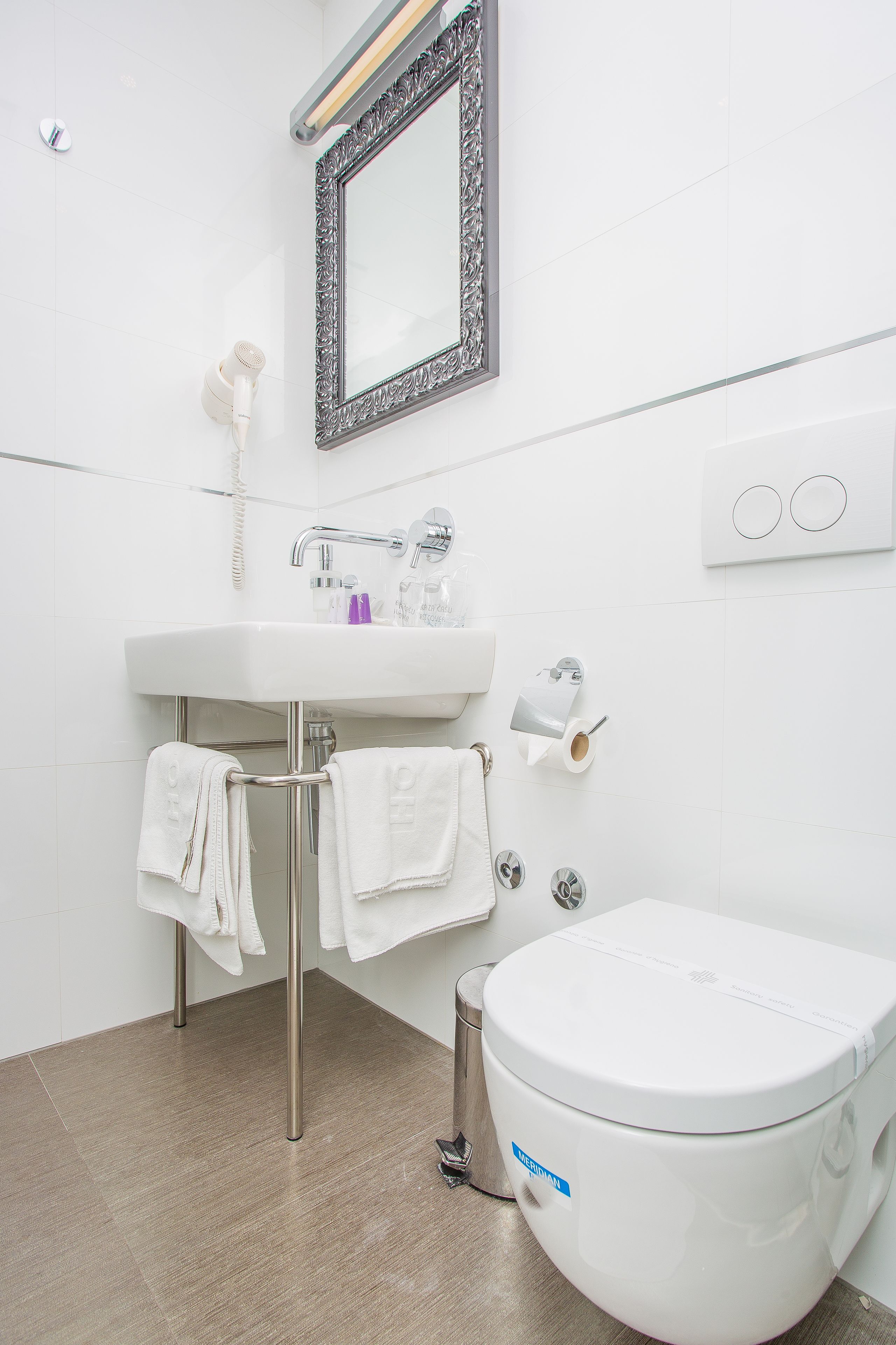 A modern bathroom with a white sink, mirror, and toilet. There are towels hanging on a metal rack, and a toilet paper holder is visible.