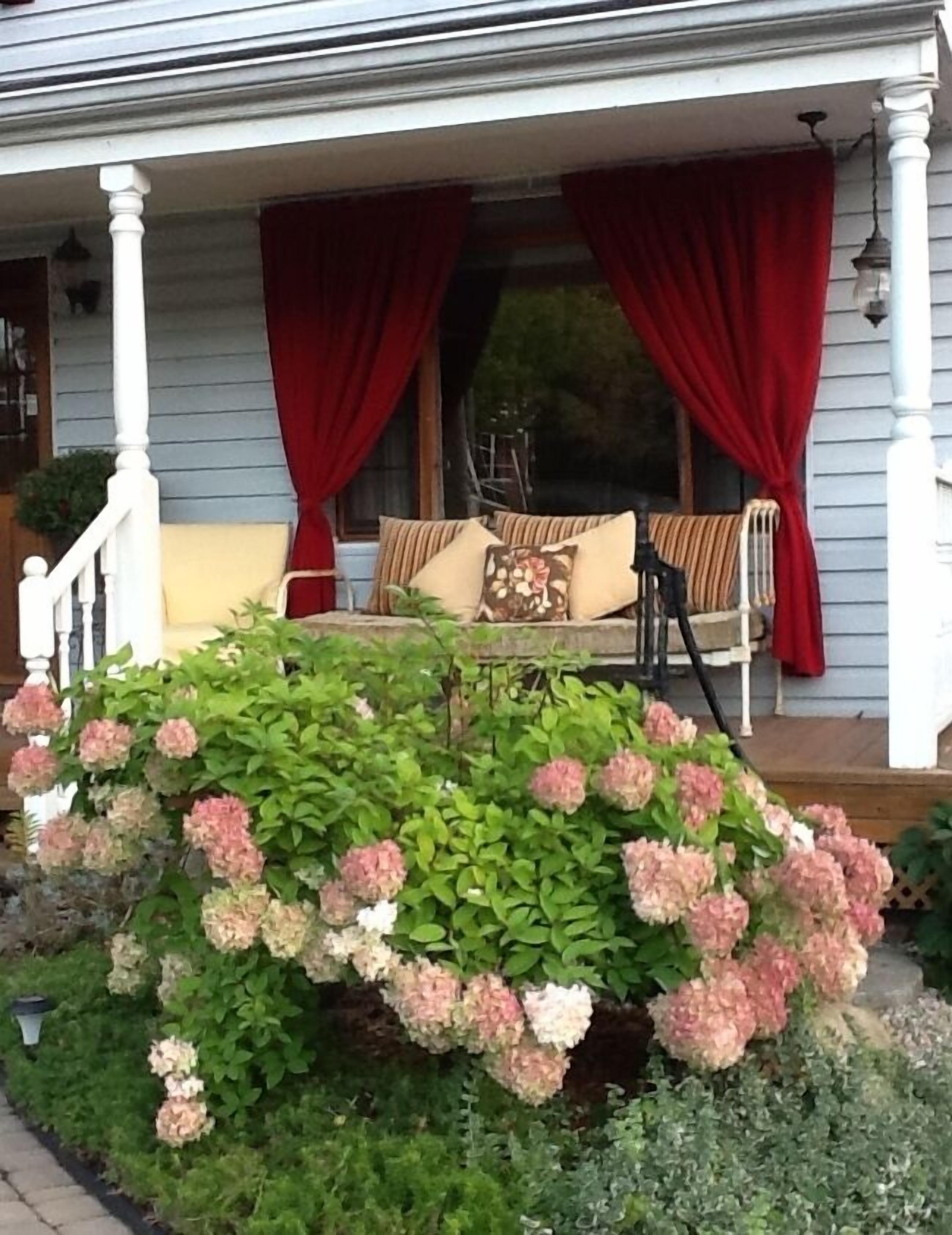 A porch with a bench, red curtains, and a flower bed with pink hydrangeas.