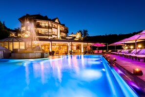 A large indoor swimming pool with a glass enclosure, a multi-story building with outdoor seating, and a well-lit patio area. - Romantischer Winkel RoLigio & Wellness Resort (Bad Sachsa)