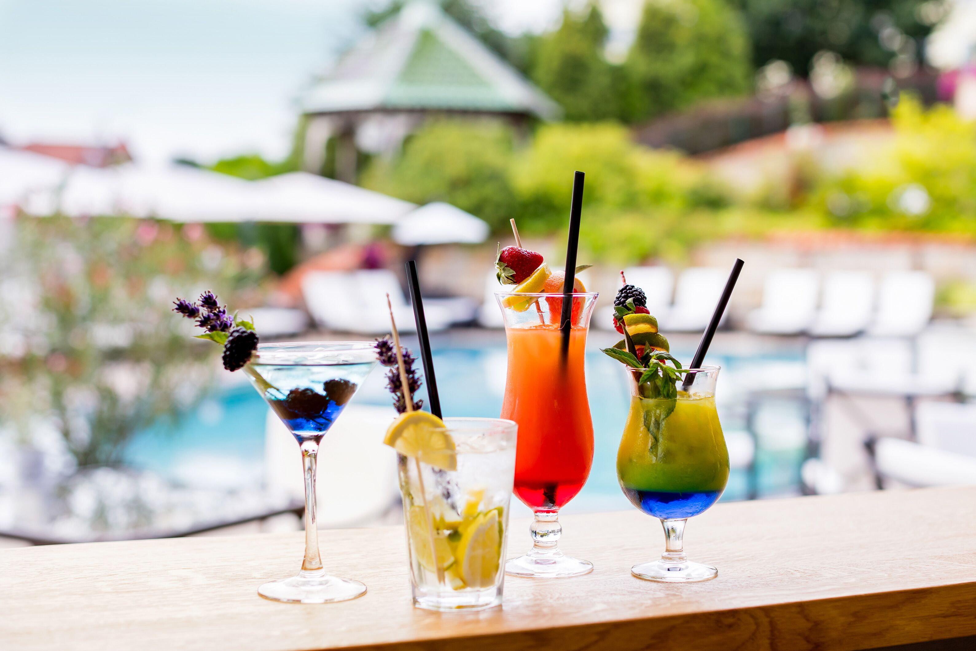 A row of four colorful cocktails on a wooden bar with a pool in the background.