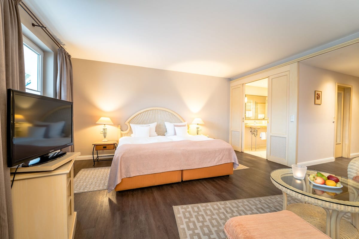 A hotel room with a bed, a TV, a bedside table with a lamp, a glass table with fruit, and a bathroom visible through an open door.
