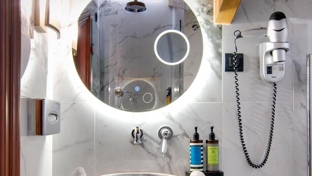 A bathroom with a copper sink, a round mirror, a handheld showerhead, and a hairdryer mounted on the wall. - Archontariki Boutique Hotel (Ioannina)