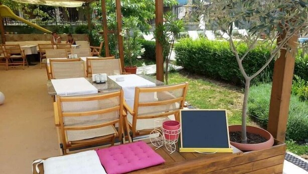 Un espacio al aire libre con mesas y sillas de madera, un cojín rosa, una pizarra y una planta en maceta.