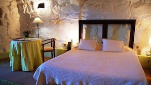 A bedroom with a large bed, a small round table, and a chair. The room has a stone wall and a lamp on the table.