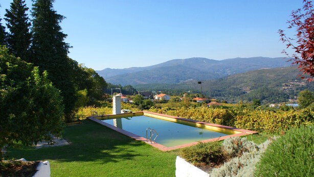 A swimming pool surrounded by greenery, with a view of distant mountains and houses. - Quinta Da Calcada (Melgaço)