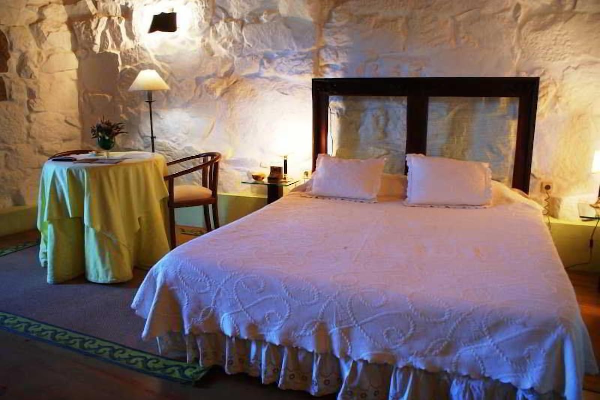 Ein Schlafzimmer mit einem großen Bett, einem kleinen runden Tisch und einem Stuhl. Der Raum hat eine Steinwand und eine Lampe auf dem Tisch.
