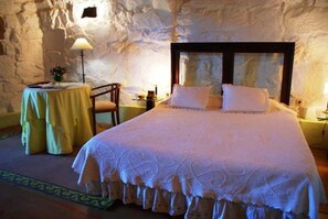 Ein Schlafzimmer mit einem großen Bett, einem kleinen runden Tisch und einem Stuhl. Der Raum hat eine Steinwand und eine Lampe auf dem Tisch.