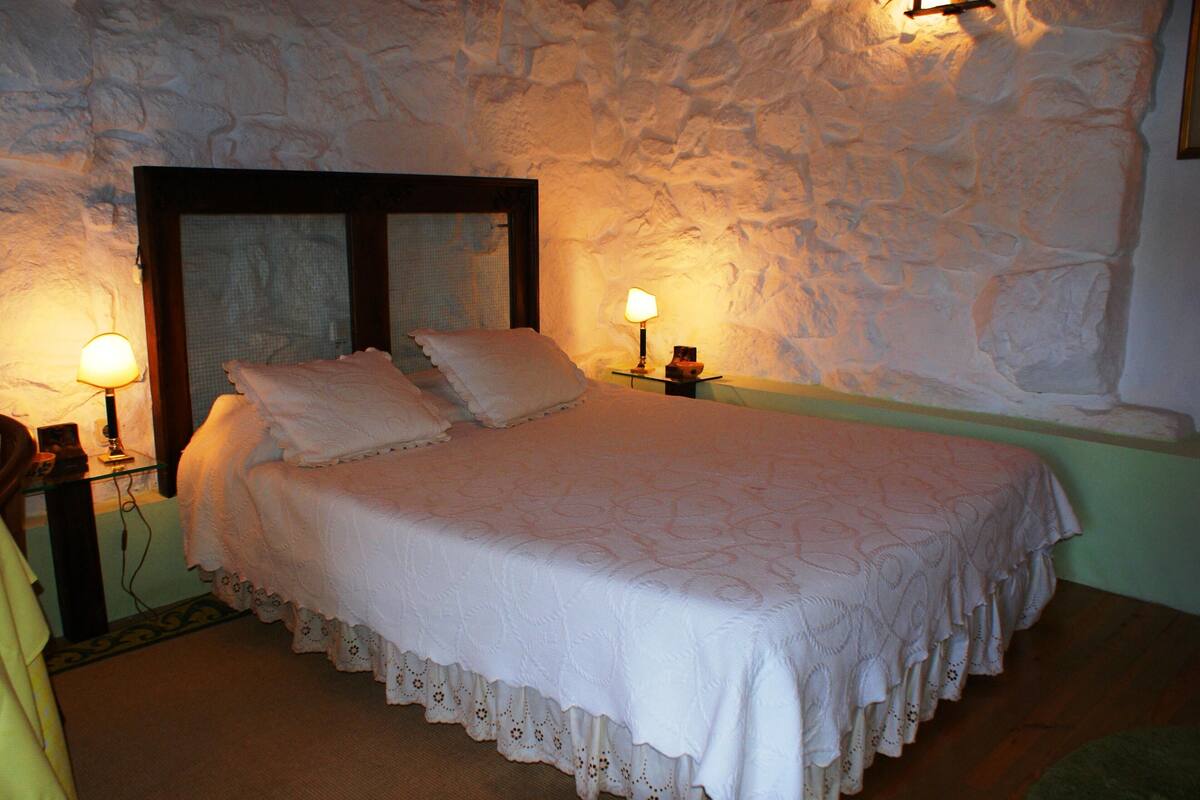 Una cama con una colcha blanca y almohadas, dos lámparas de noche, una mesita de noche y una pared de piedra.