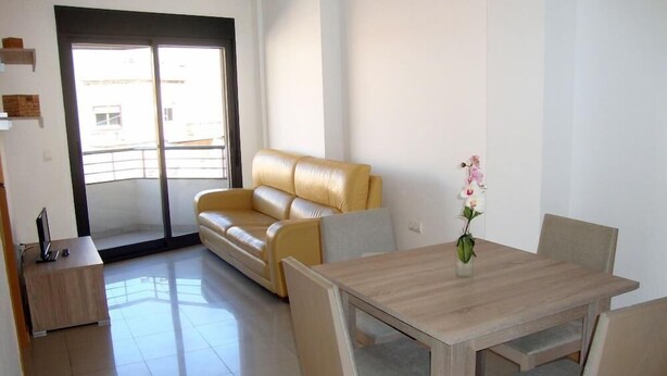 A modern living room with a sofa, a dining table with chairs, a small side table, and a window with a view of buildings. - Benicarlo 3000 (Benicarlo)