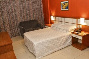 A hotel room with a bed, a chair, a nightstand with a phone, and a wall-mounted lamp.