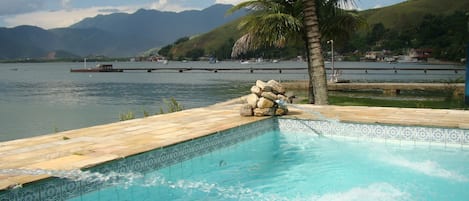 A swimming pool with a tiled edge, a palm tree, and a view of a lake with boats and mountains in the background.