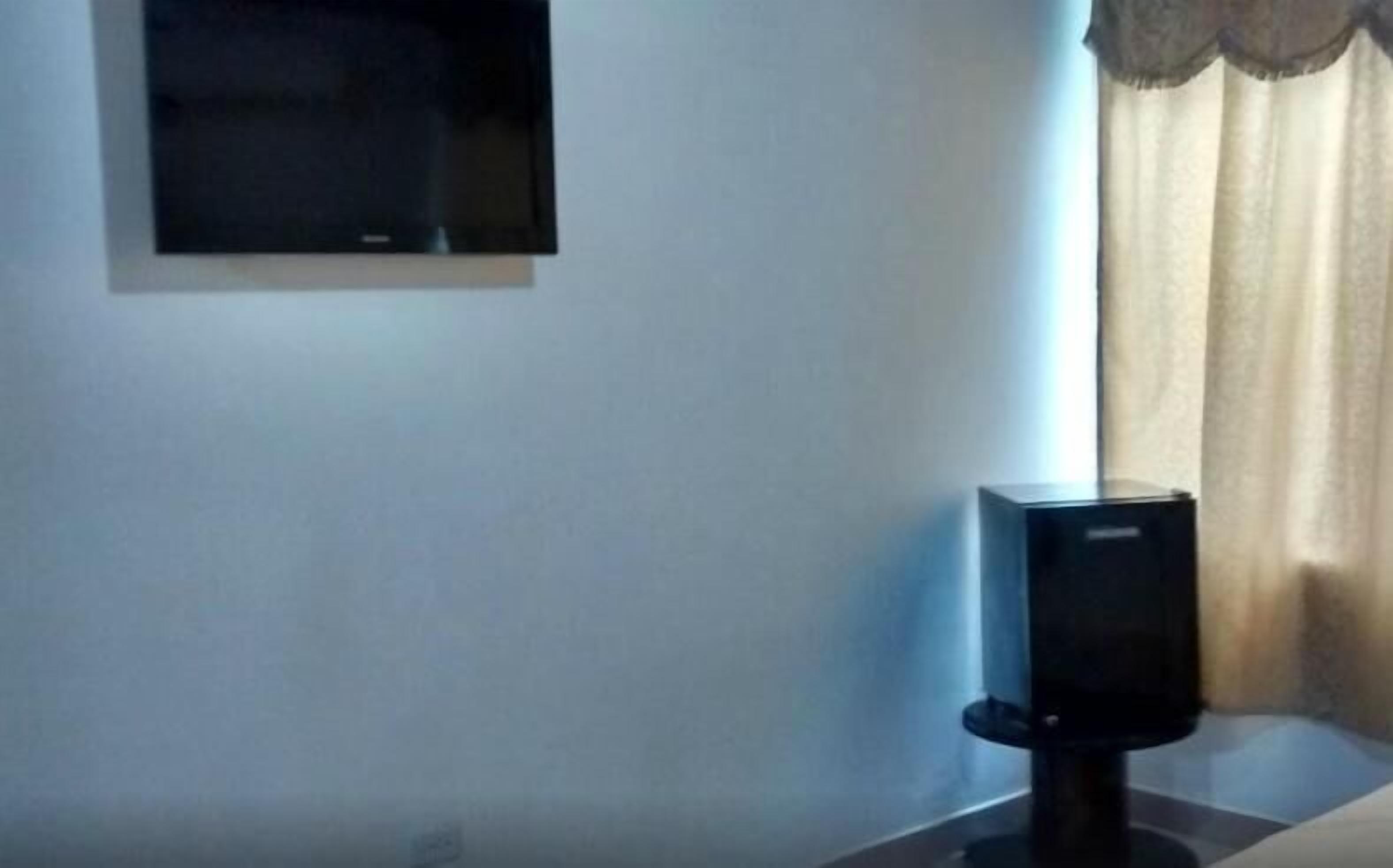 A room with a flat-screen TV mounted on the wall, a black speaker on a table, and a window with curtains.