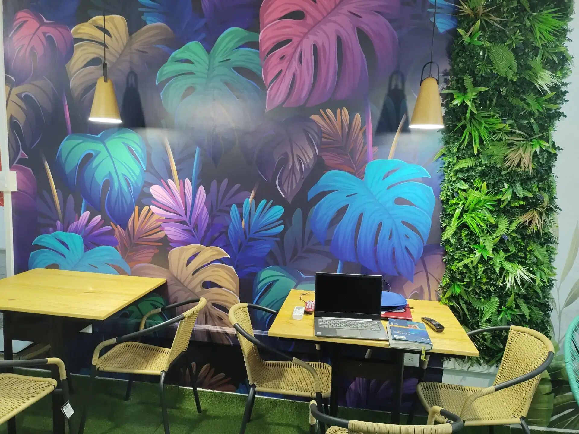 A room with a vibrant tropical leaf mural, a green wall with foliage, wicker chairs, and a table with a laptop.
