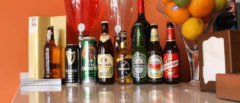 Variedad de botellas y vasos sobre una mesa, incluyendo cerveza y vino, con un frutero al fondo.