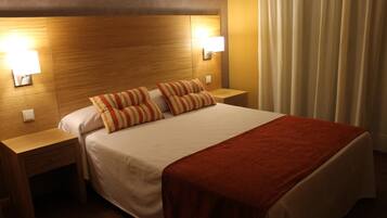 Une chambre d’hôtel avec une tête de lit en bois, deux lampes de chevet, un lit avec des oreillers à rayures, un plaid rouge et un rideau à la fenêtre.