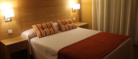 Une chambre d’hôtel avec une tête de lit en bois, deux lampes de chevet, un lit avec des oreillers à rayures, un plaid rouge et un rideau à la fenêtre.