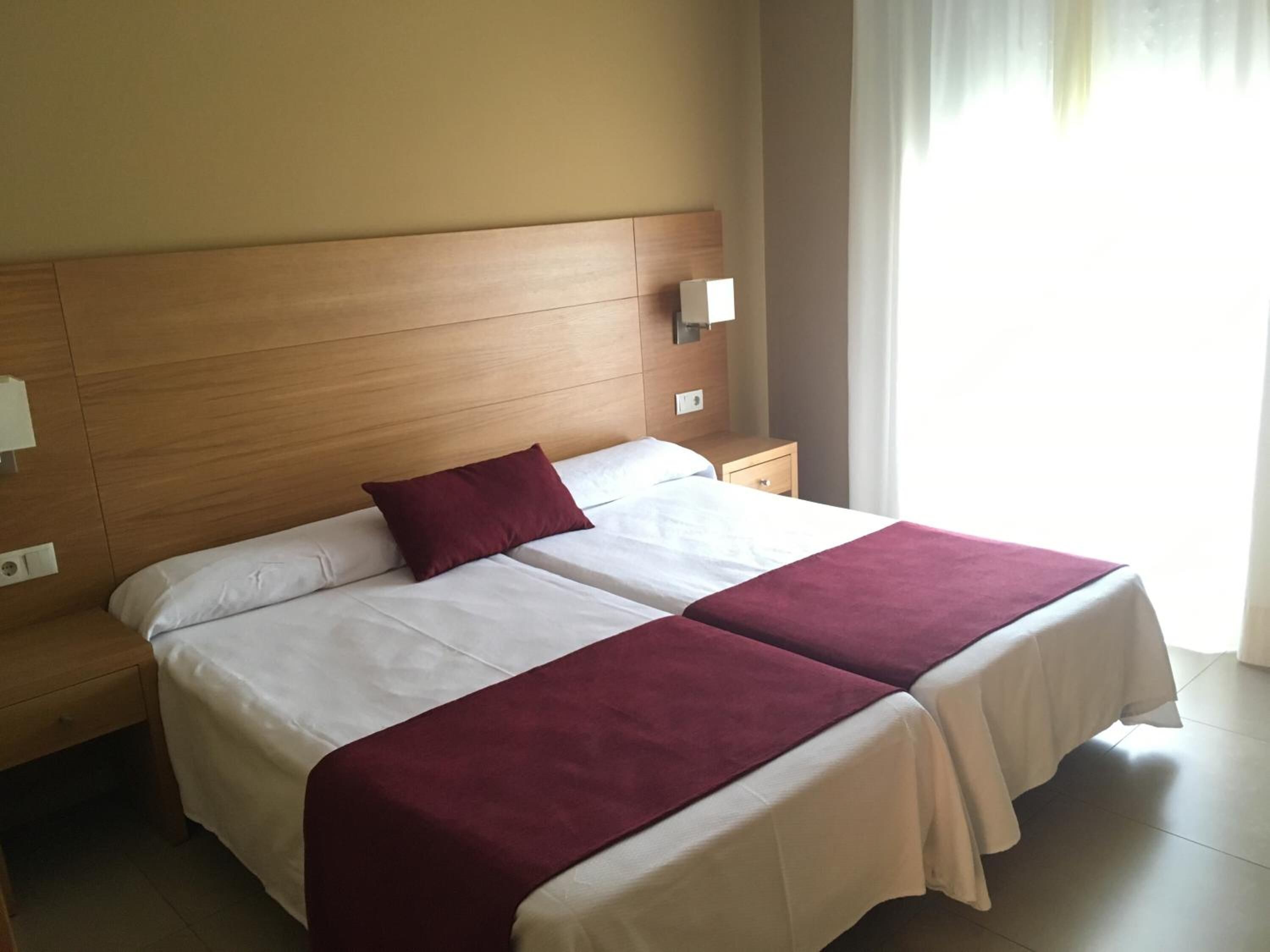 Un lit double avec une literie blanche et bordeaux, une tête de lit en bois, une table de chevet avec une lampe et une fenêtre avec des rideaux.