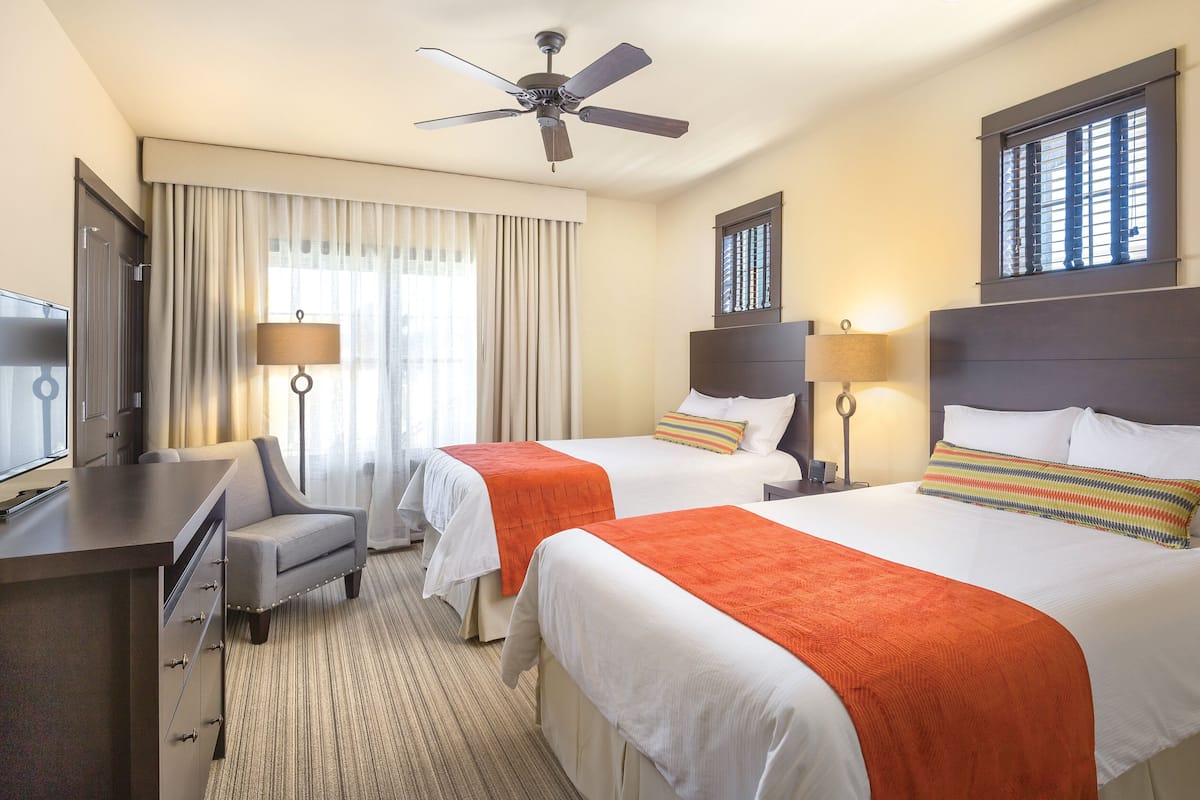 A hotel room with two beds, a desk, a chair, a ceiling fan, and a window with blinds.