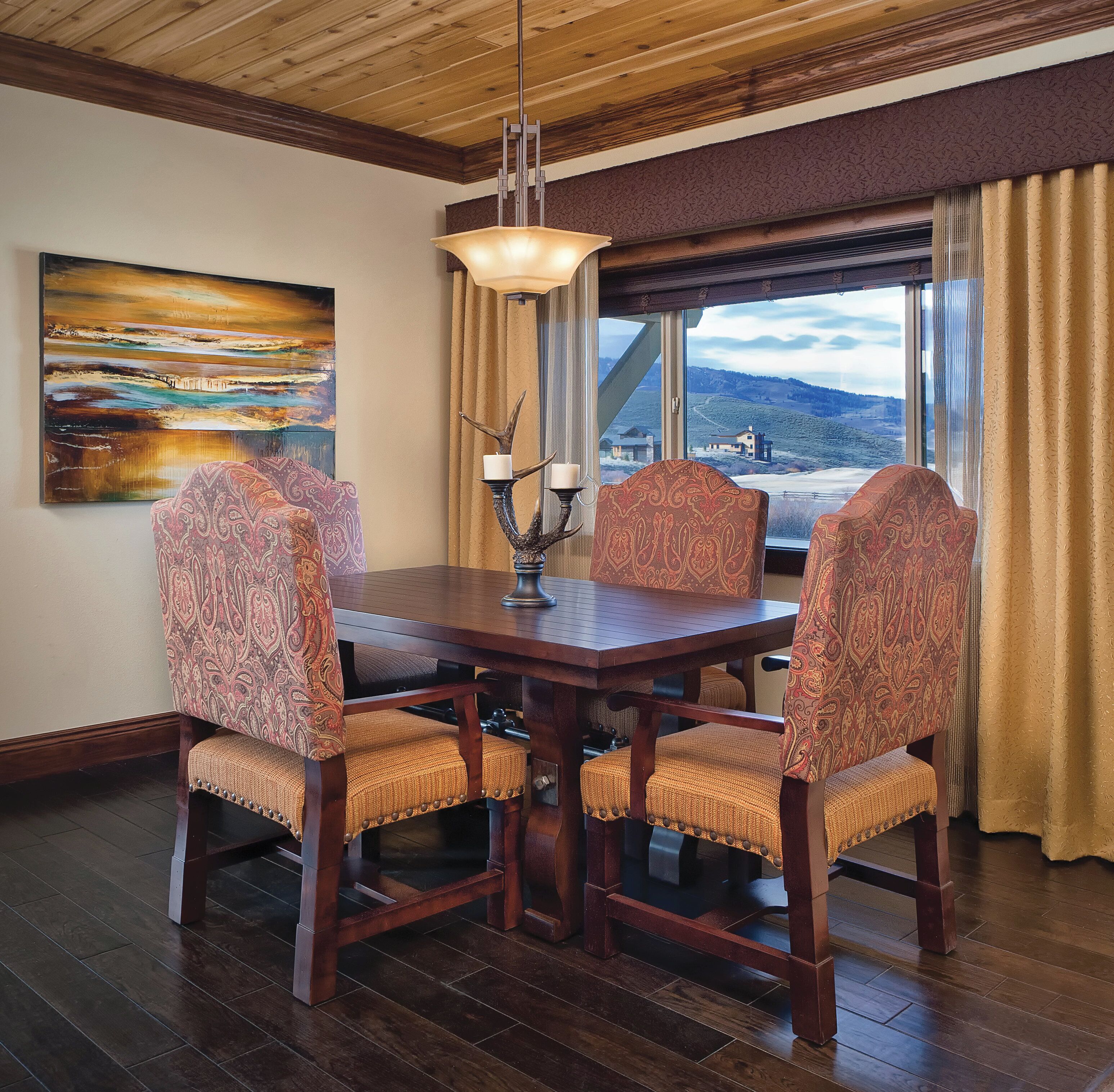 A dining area with a wooden table and patterned chairs, a painting on the wall, and a view of a landscape through large windows.