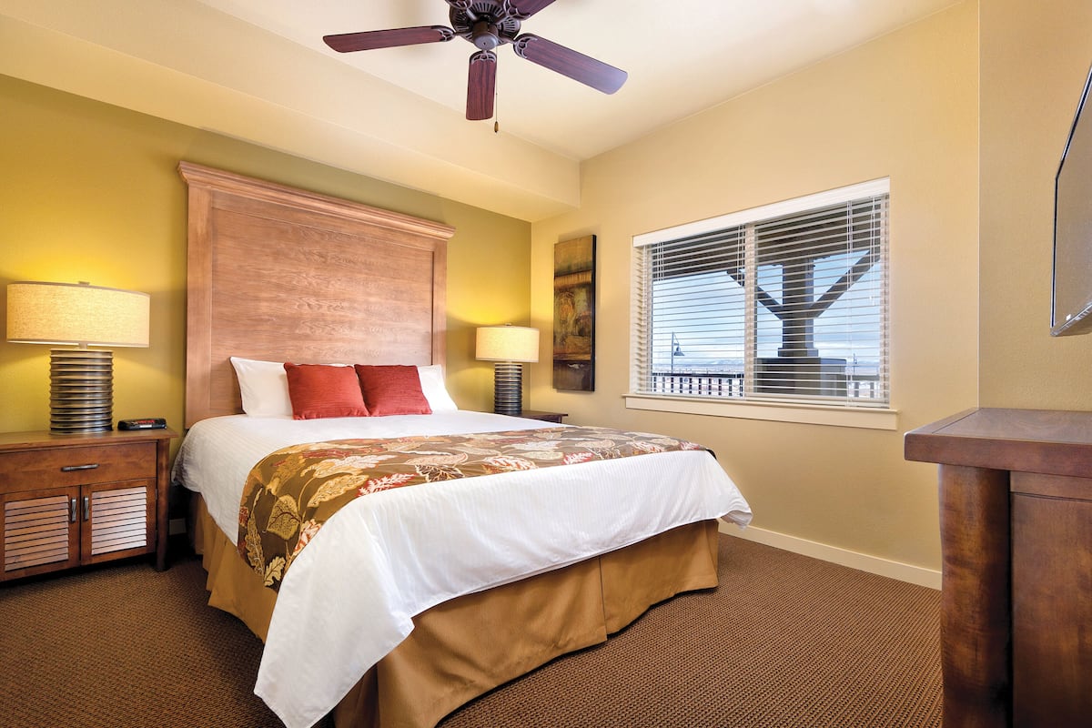 A bedroom with a large bed, two bedside tables with lamps, a ceiling fan, a window with blinds, and a TV mounted on the wall.