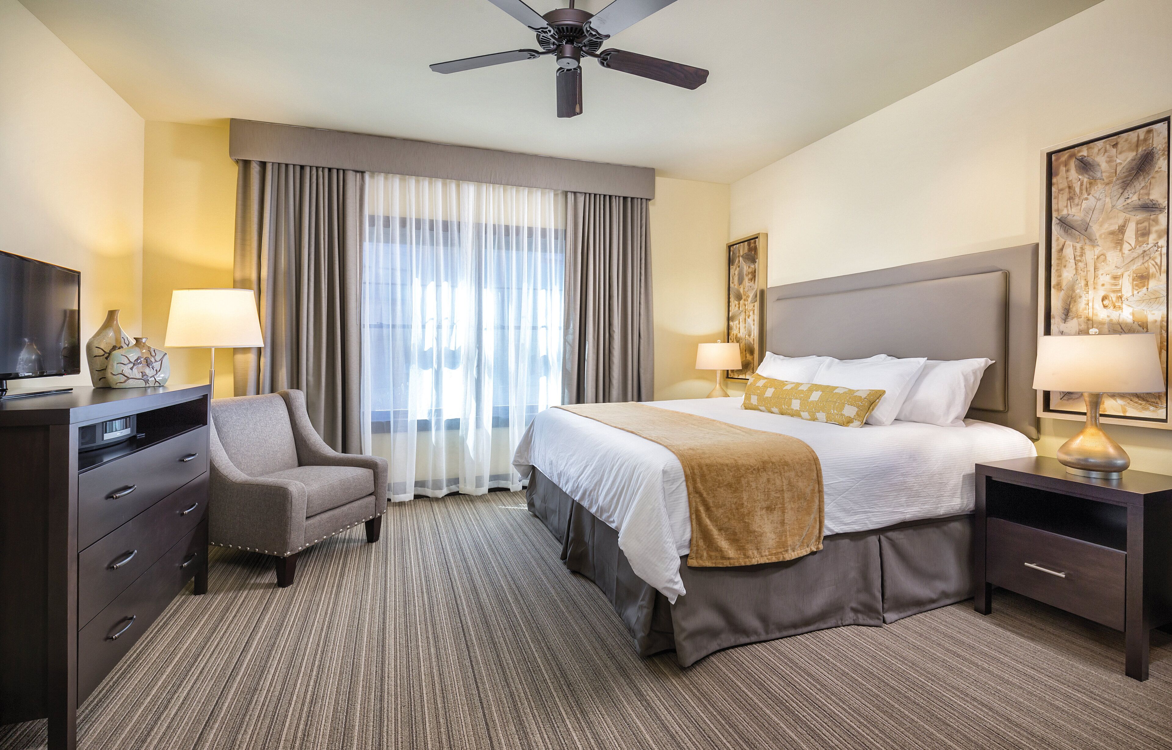 Quarto de hotel com cama, escrivaninha, cadeira, televisão e ventoinha no teto.