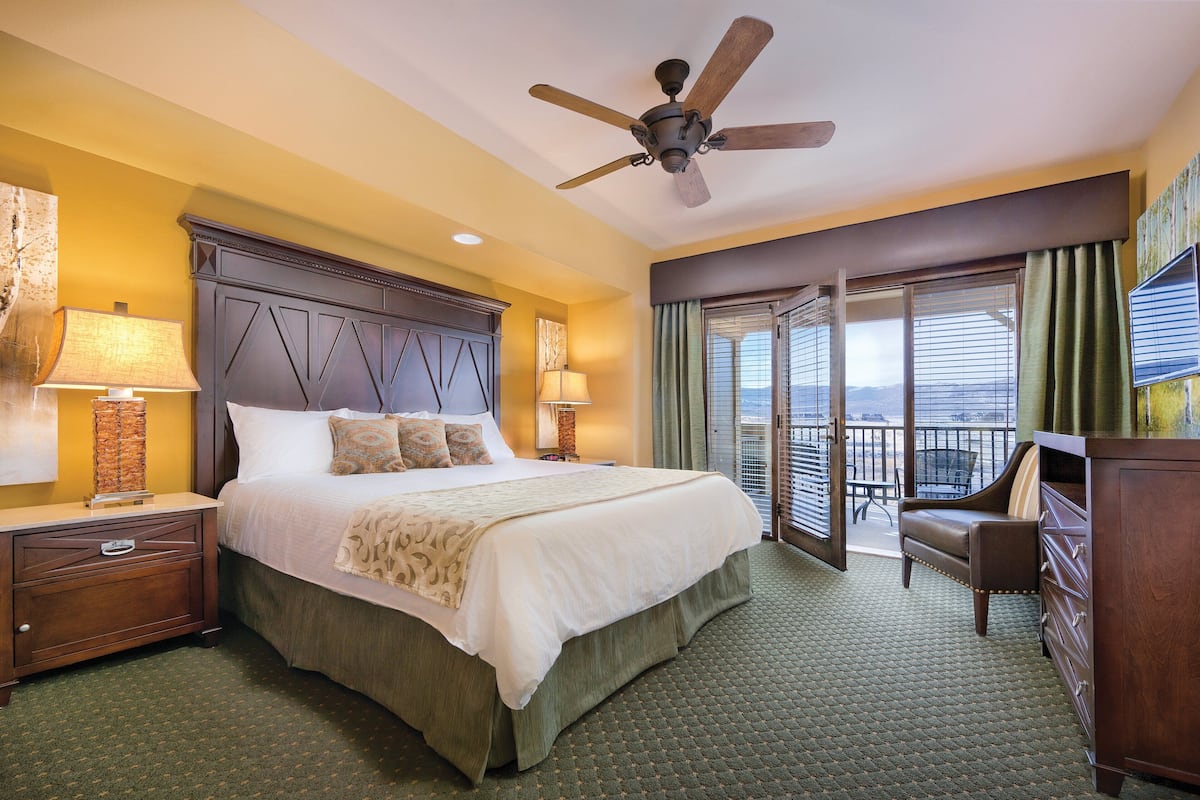 A hotel room with a large bed, a ceiling fan, a nightstand with a lamp, a dresser, and a balcony with a chair.
