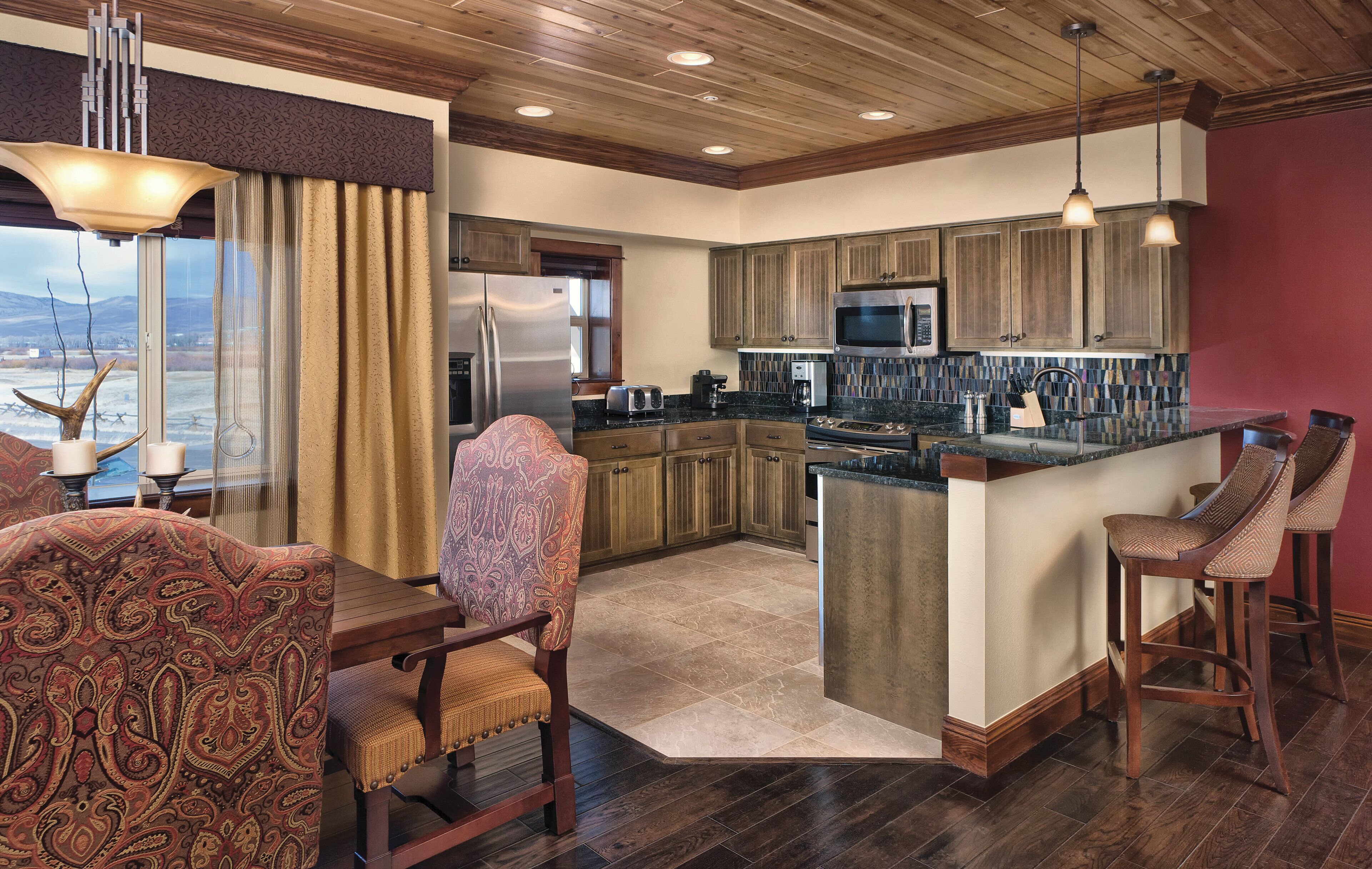A modern kitchen with wooden cabinets, a central island, bar stools, and a dining area with patterned chairs.